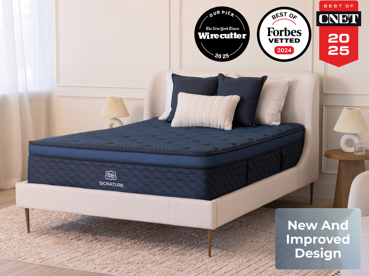 The Brooklyn Bedding Signature Hybrid mattress styled on a cream upholstered bed in a bright bedroom with award badges above and a New And Improved Design text box.